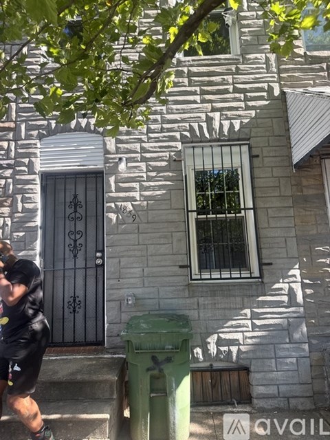 A man in a black shirt and shorts is running on a sidewalk past a green trash can and a house with a white door and window.