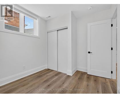 A room with white walls and wooden flooring, with a window showing a brick building outside.