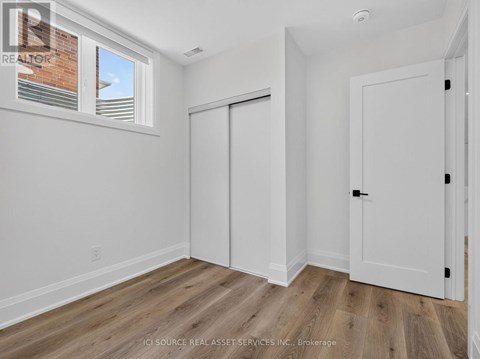 A room with white walls and wooden flooring, with a window showing a brick building outside.