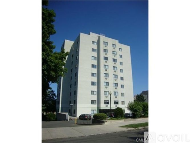 A tall white building with many windows is surrounded by trees and a car is parked in front.