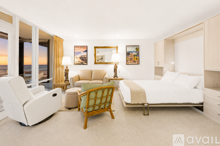 A well-lit bedroom with a large bed, a chair, a sofa, and a view of the beach through the window.