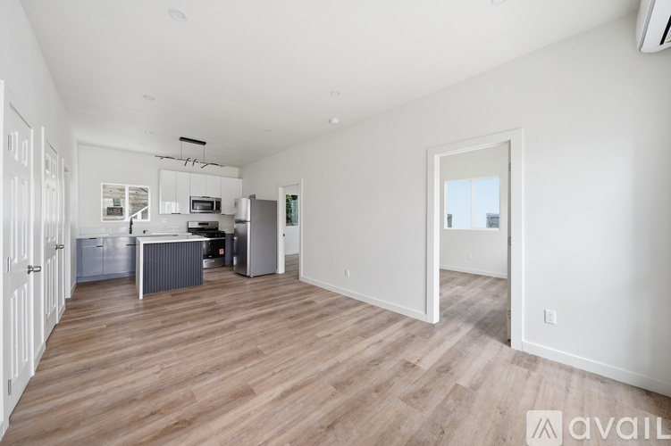 A spacious room with wooden flooring and white walls, leading to a kitchen area.