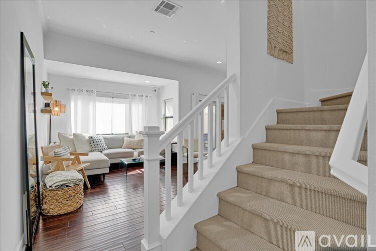 A staircase with beige carpeted steps and white railings leads to a bright living room with a white couch and a window with white curtains.