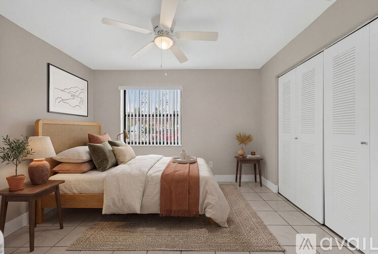 A bedroom with a bed, a nightstand, a table, a potted plant, and a window with a view of a balcony.