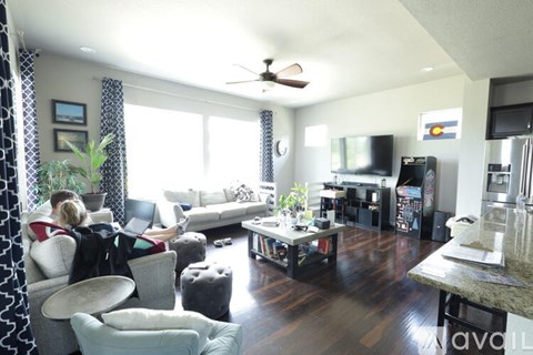 A woman is sitting on a couch in a living room with a television, a fan, and a kitchen area in the background.
