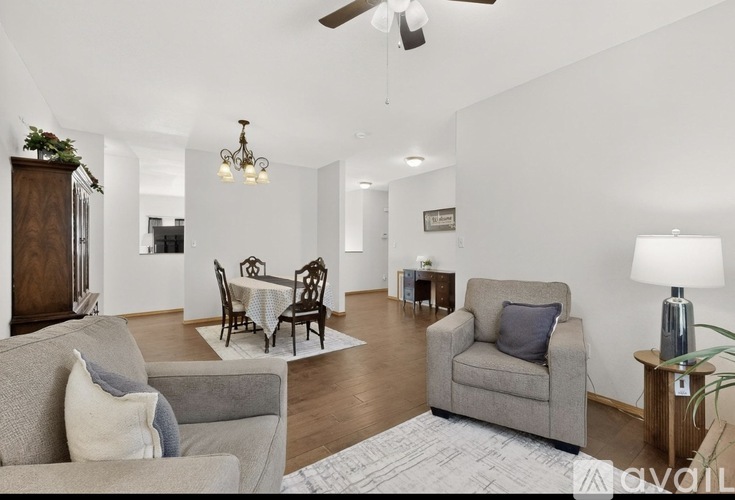 A living room with a grey couch, a grey armchair, a dining table with chairs, a ceiling fan, and a plant.