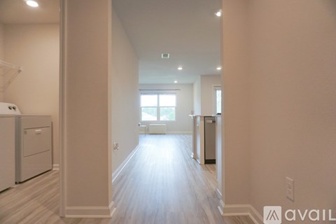 A spacious kitchen with white cabinets and a refrigerator, leading to a bright room with a window.