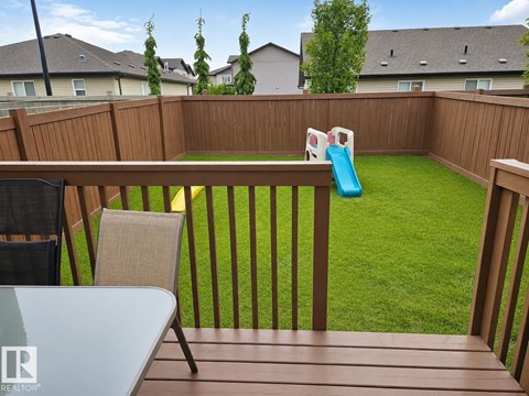 A backyard with a wooden deck, a table, two chairs, a slide, and a green lawn.