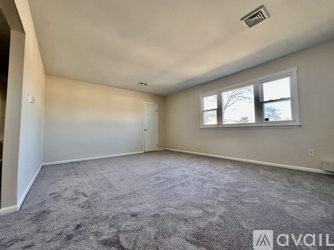 A spacious room with a carpeted floor, a window with a view of trees, and a ceiling fan.