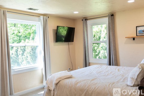 A bedroom with a bed, a window with curtains, and a flat-screen TV mounted on the wall.