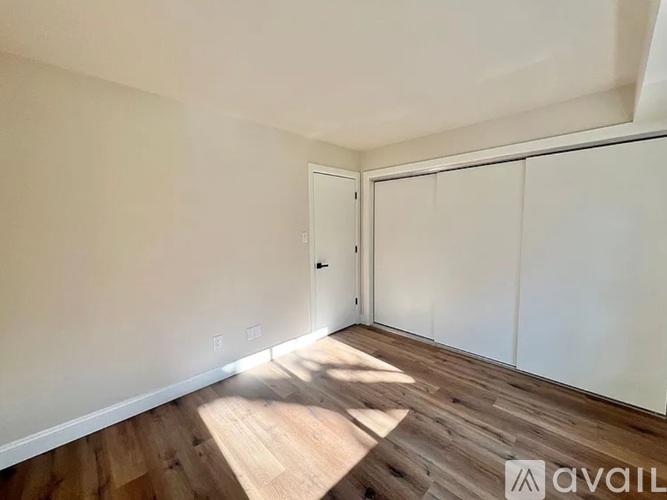 A room with wooden flooring and white walls, with a door and a window letting in sunlight.