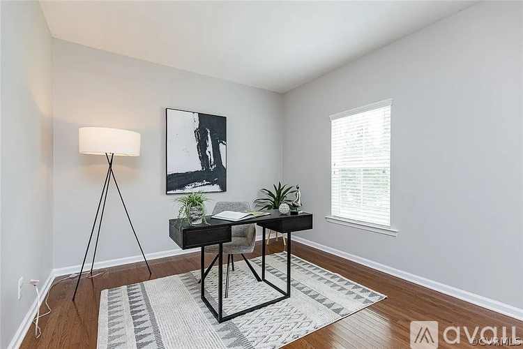 A room with a black and white photo on the wall, a small table with a lamp and two plants, and a window with blinds.