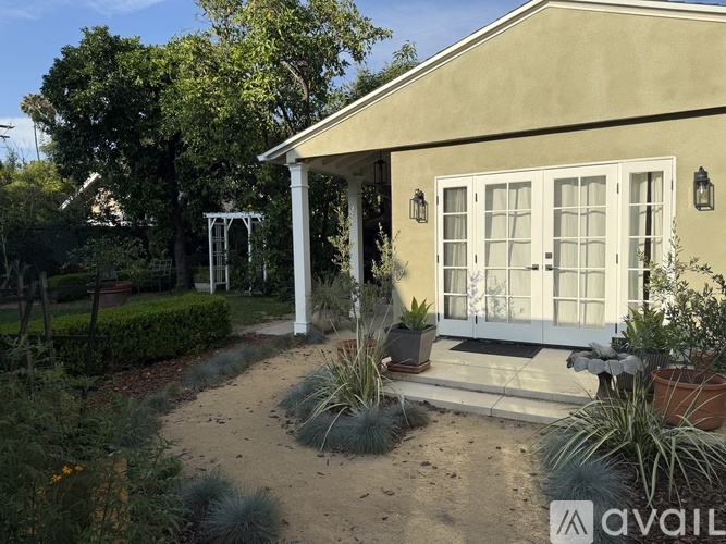 A house with a white door and windows is surrounded by plants and trees.