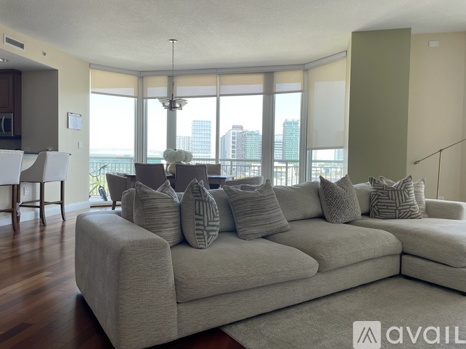 A large beige couch with multiple pillows sits in a spacious living room with a view of the city.