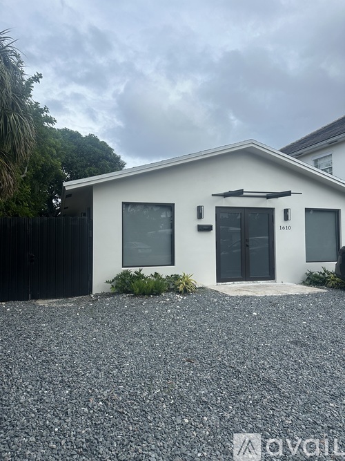 A white house with a black door and windows is surrounded by a gravel driveway.