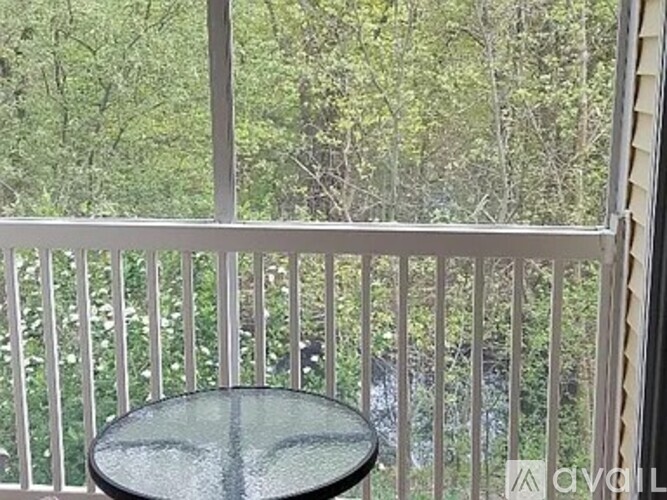 A glass table is on a white balcony with a view of trees.