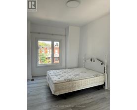 A white bed with a floral pattern mattress sits in a room with a window showing a view of a red brick building.