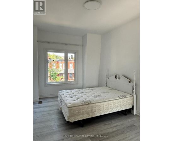 A white bed with a floral pattern mattress sits in a room with a window showing a view of a red brick building.