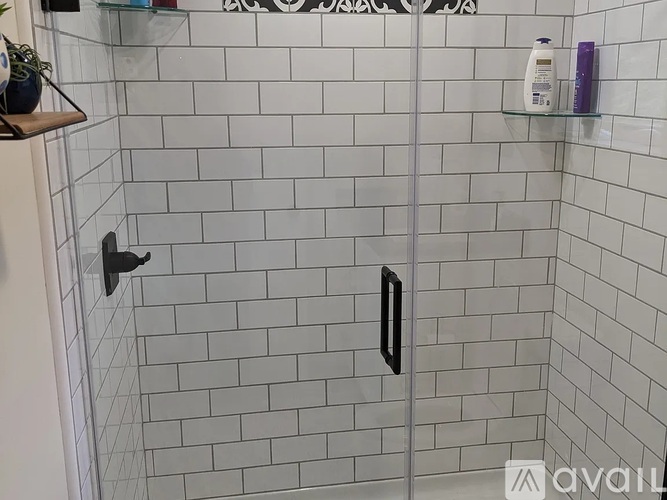 A white tiled shower with a black handle and a shelf above it.