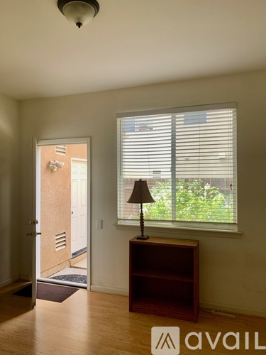 A room with a wooden floor, a window with blinds, a lamp on a wooden stand, and a door leading to another room.