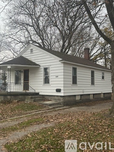 A white house with a black roof and a chimney is surrounded by trees and fallen leaves.