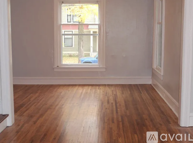 A room with wooden flooring and a window showing a blue car and a building outside.