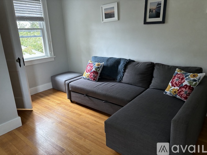 A grey couch with a floral pillow sits in a room with wooden floors and a window.
