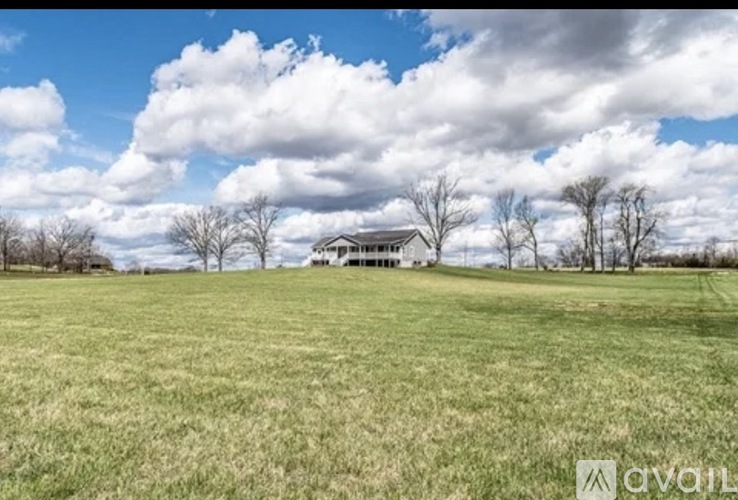 A house is situated in a field with trees and clouds in the background.