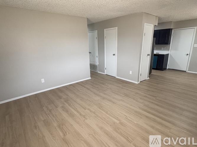 A room with wooden flooring and white walls, with a door on the left and a kitchen area on the right.