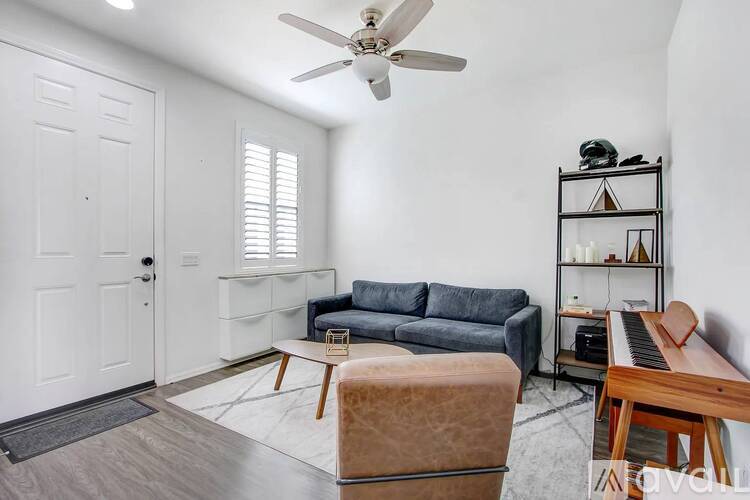 A living room with a grey couch, a brown ottoman, a piano, and a ceiling fan.