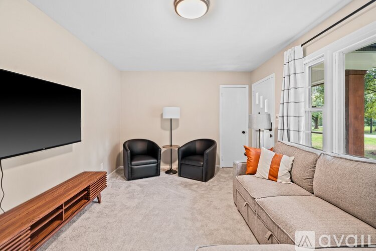 A living room with a flat screen TV, two black chairs, a grey sofa, and a window with a view of the outdoors.