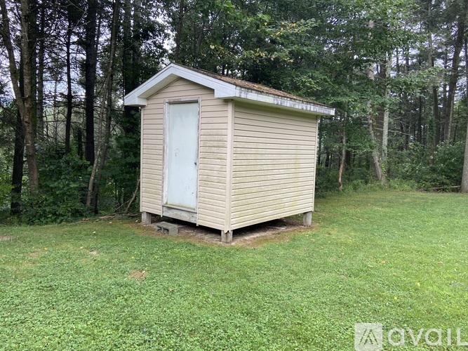 A small beige shed with a white door is situated in a grassy area with trees in the background.