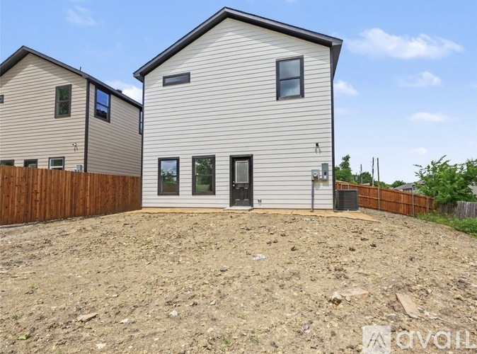 A modern house with a grey exterior and a black door is situated in a sandy area with a wooden fence.