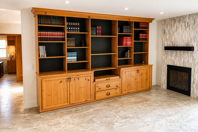 A large wooden bookshelf with books and decorative items is in the center of a spacious room with a fireplace on the right side.