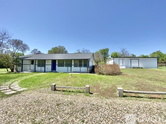 A house with a white and blue exterior is surrounded by a gravel driveway and a grassy area with a small bush.