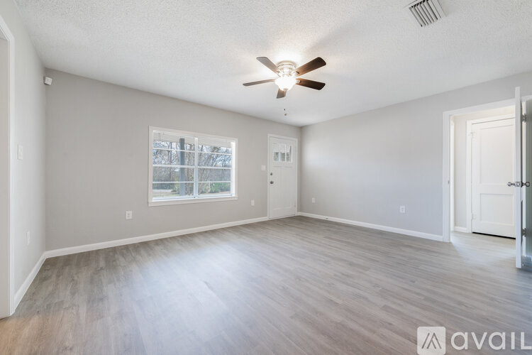 A spacious room with a ceiling fan and light, hardwood floors, and a window with a view of trees outside.