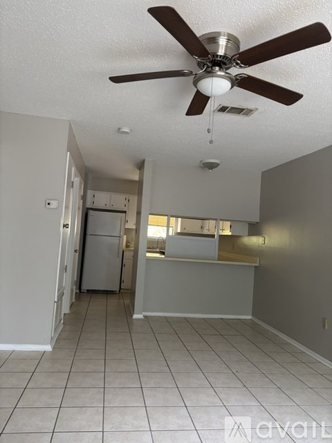 A room with a ceiling fan and a kitchen area in the background.