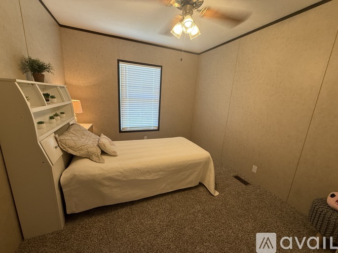 A small bedroom with a bed, a window with blinds, a ceiling fan, and a small shelf with a plant on it.