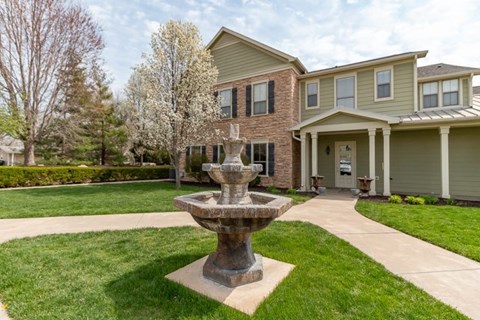 A stone fountain is in the middle of a grassy area in front of a house.