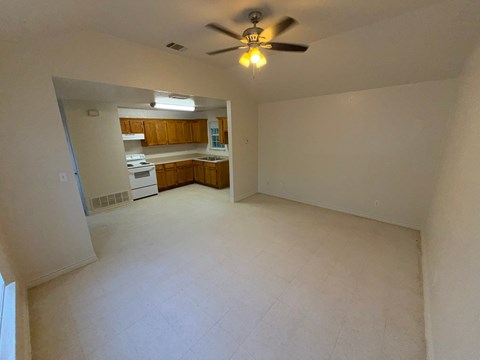 A room with a ceiling fan and a kitchen area in the background.