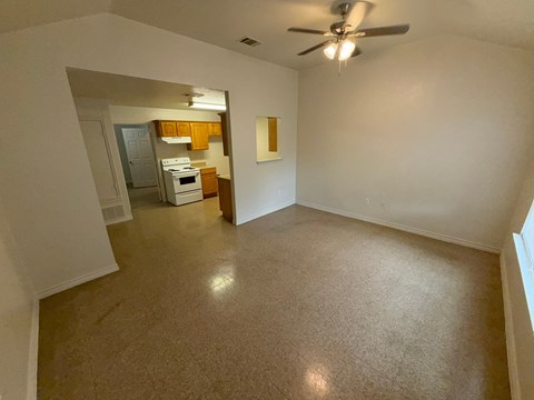 A room with a ceiling fan and a kitchen area in the background.