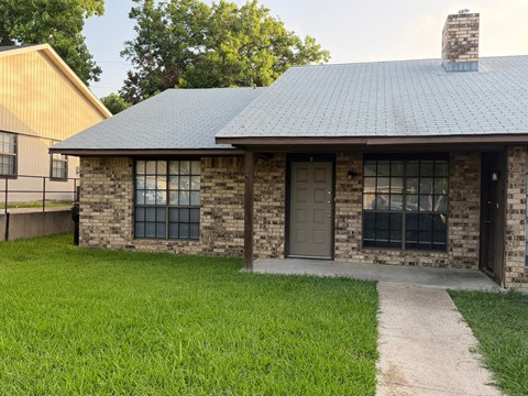 A small house with a grey door and windows is surrounded by a grassy area.