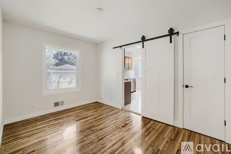 A room with wooden flooring and white walls, a window with a view of trees, and a door on the right.