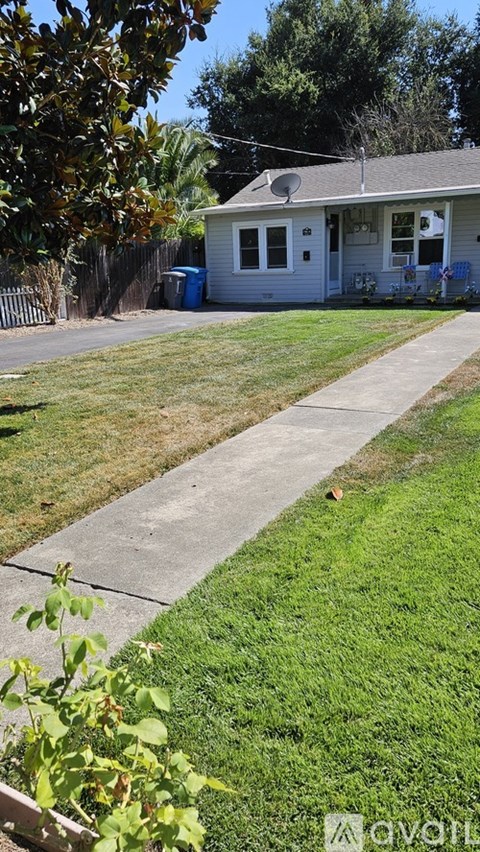 A house with a grey roof and a blue door is surrounded by a green lawn and a concrete pathway.