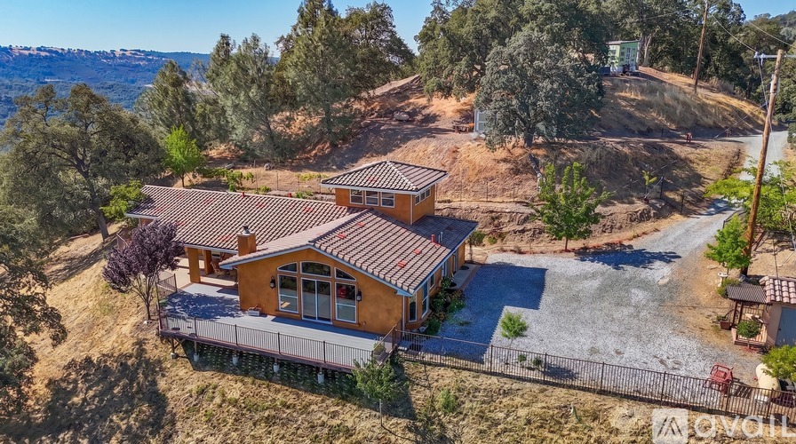 A house with a brown roof and a balcony is surrounded by trees and a hillside.