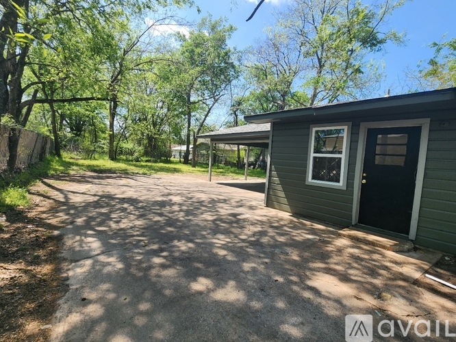 A small house with a black door is surrounded by trees and has a gravel driveway.