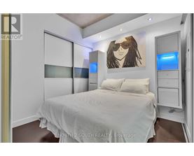 A bedroom with a bed, a dresser, and a painting of a woman with sunglasses on the wall.