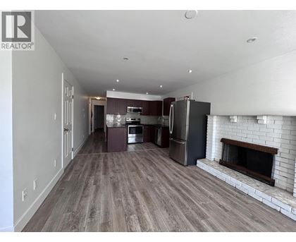 A large, empty living room with a fireplace and a kitchen area in the background.