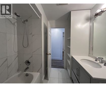 A bathroom with a white tub and sink, a shower with a hose, and a door leading to another room.