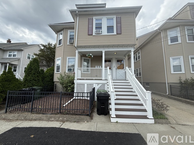 A two-story house with a front porch and a black trash bin on the sidewalk.